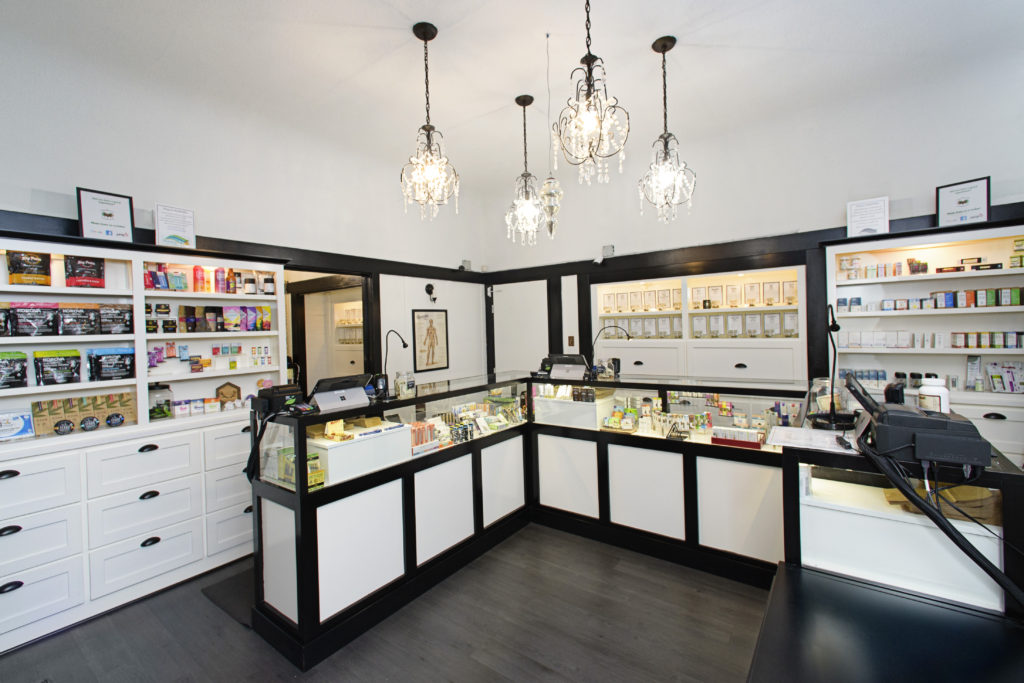 A well-lit modern store interior featuring three elegant chandeliers hanging from the ceiling exudes a therapeutic ambiance. Display counters with glass showcases are filled with various products, while shelves on the walls behind the counters hold an assortment of alternative goods. CA Norml