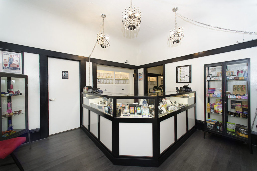 A well-lit interior of a store with a black and white color theme. The store features glass display cases filled with various therapeutic products, hanging decorative lights, and posters on the walls. There is a door with a restroom sign and an alternative seating area in the foreground. CA Norml