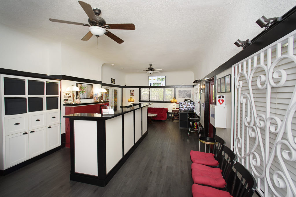 A spacious room with white and black decor, including a sweeping counter with a white front and black trim. The red cushioned seating along the right wall offers therapeutic comfort, facing an ornate white partition. A ceiling fan is suspended from the white ceiling, while various artworks adorn the walls. CA Norml