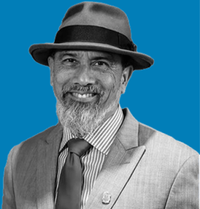 A man with a greying beard and mustache, wearing a fedora hat and a light-colored suit with a striped shirt and tie, smiles at the camera. The background is solid blue. He represents Armada Law Corp. — Ca NORML Legal Committee member. CA Norml