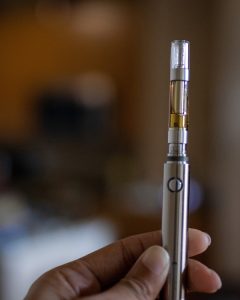 A hand is holding a sleek silver vape pen with a clear cartridge partially filled with a brown liquid. The background is blurred, suggesting an indoor setting. CA Norml