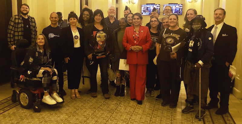 A diverse group of people posing in a warmly lit room. Some are standing, while one person is in a wheelchair. Dressed in both casual and formal attire, they gather cheerfully, reflecting on 2024 accomplishments while eagerly discussing plans for 2025. CA Norml