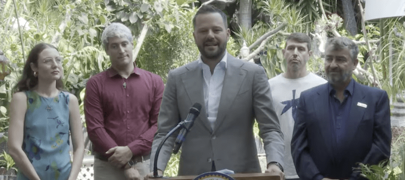 A man in a suit speaks at a podium outdoors, flanked by two men and two women, highlighting 2024 accomplishments. The podium displays a seal that reads "State Assembly," as lush greenery forms the backdrop, hinting at future plans for 2025 with Cal NORML's commitment to progress. CA Norml