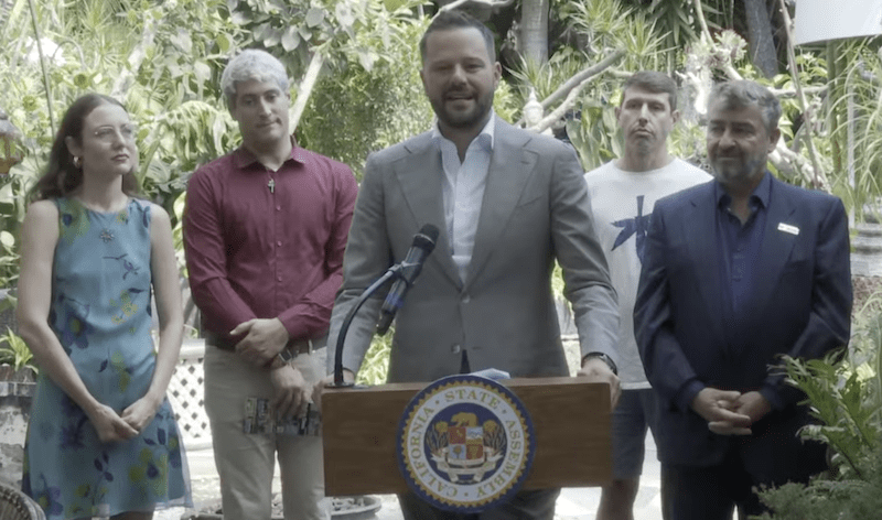 A man in a suit speaks at a podium outdoors, flanked by two men and two women, highlighting 2024 accomplishments. The podium displays a seal that reads "State Assembly," as lush greenery forms the backdrop, hinting at future plans for 2025 with Cal NORML's commitment to progress. CA Norml