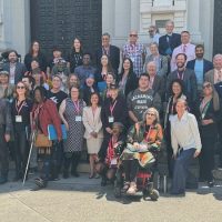 A diverse group of people, including individuals with mobility aids, stands smiling on steps in front of a grand building. Many wear lanyards, hinting at a Cal NORML conference or event gathering. The backdrop features large, decorative doors celebrating their accomplishments. CA Norml