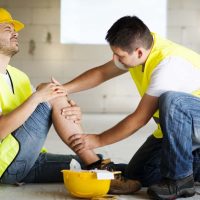 A construction worker wearing a yellow hard hat and vest sits on the ground holding his injured knee. Another worker in a yellow vest kneels beside him, offering assistance. Injured workers like him may be eligible for workers' compensation. A hard hat is placed on the ground nearby. CA Norml