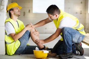 A construction worker wearing a yellow hard hat and vest sits on the ground holding his injured knee. Another worker in a yellow vest kneels beside him, offering assistance. Injured workers like him may be eligible for workers' compensation. A hard hat is placed on the ground nearby. CA Norml