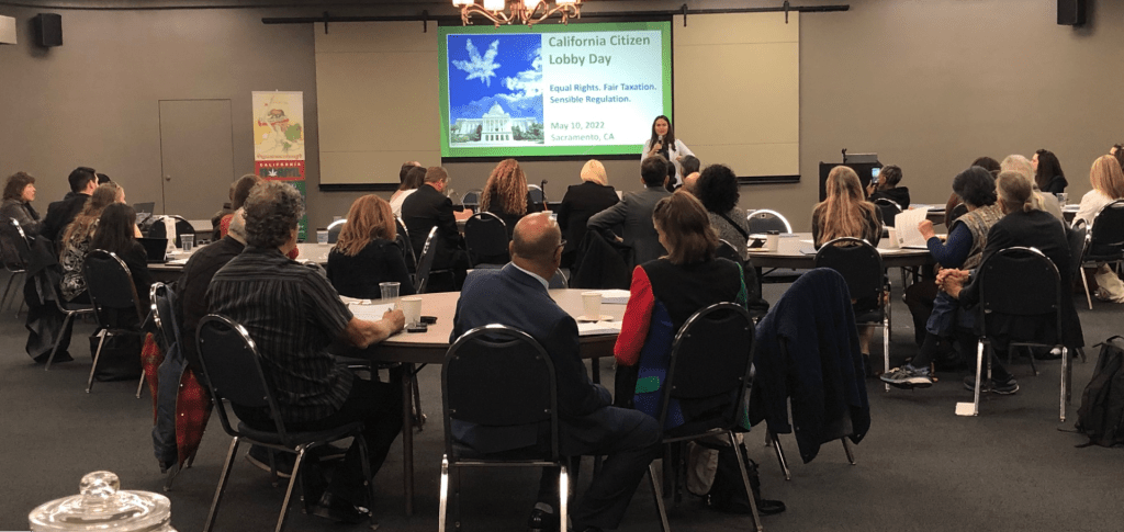 A group of people seated in a conference room listens to a speaker at the front. A large screen displays "California Citizen Lobby Day" with details and a graphic. Participants are sitting at tables, some taking notes, and a banner is visible on one side. CA Norml